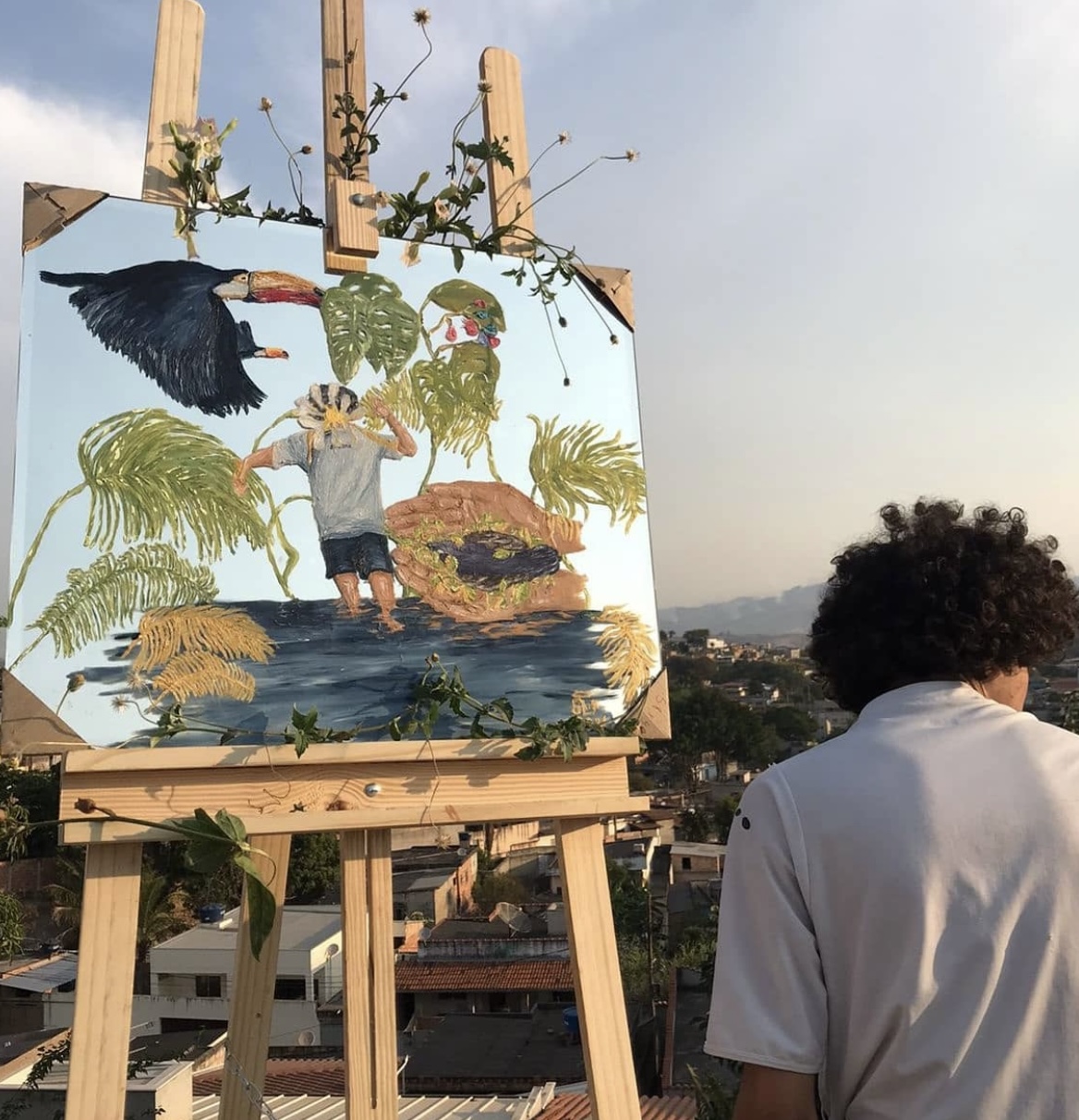 O artista de costas observando a vista ao lado da obra no cavalete
