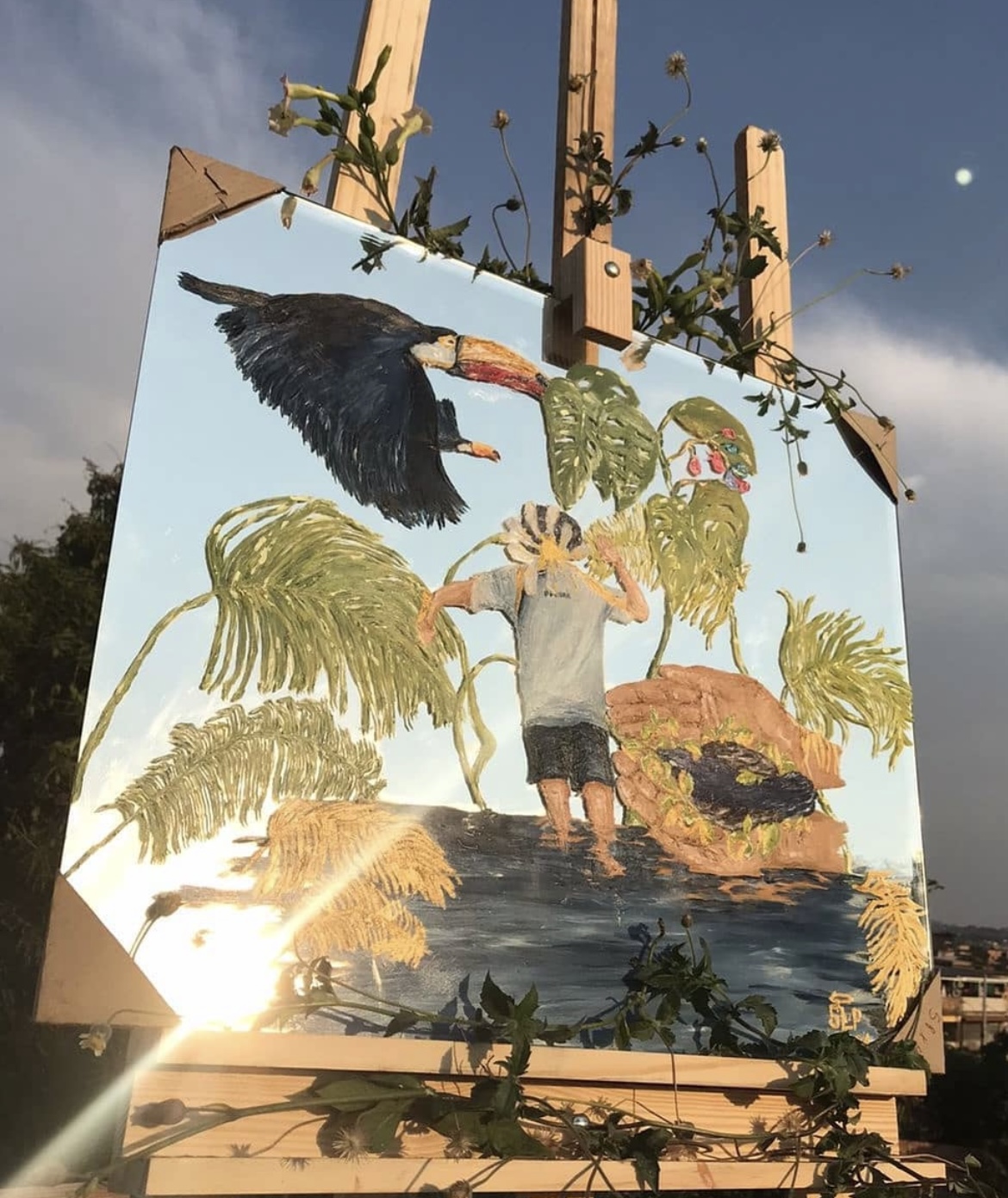 Pintura sobre espelho com tucano e plantas refletindo o céu azul, apoiada em um cavalete ao ar livre